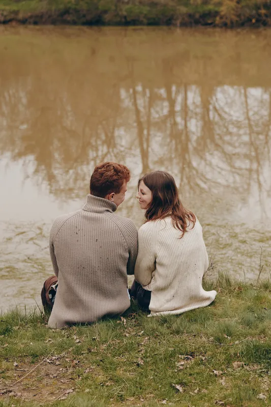 A couple sits side by side on the grass by a calm lake, wearing cozy sweaters, sharing a quiet moment, and enjoying a gentle autumn scene in a serene outdoor park setting.