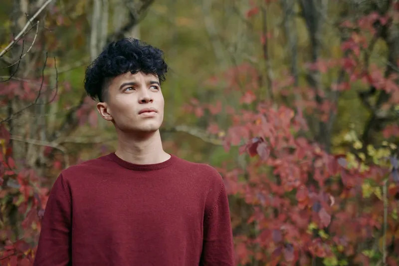 A portrait of a young man in a maroon sweater among autumn trees, showing natural posture, honest facial expression and intimate feel that highlights authenticity (?????????????) and ???????.