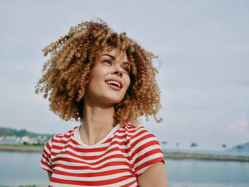 Portrait of a cheerful woman with curly hair wearing a red and white striped tee, standing outdoors in bright daylight with a calm natural backdrop.