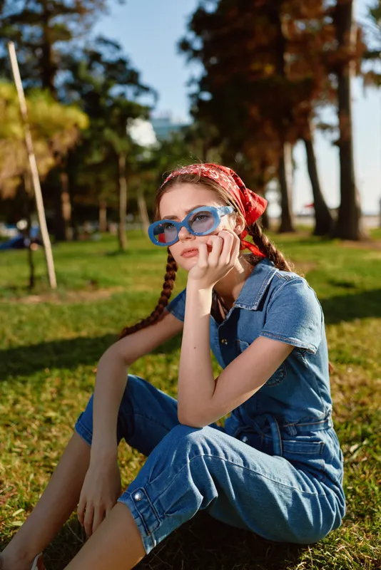 Lifestyle woman in modern boho-western denim street style wearing blue sunglasses and red headscarf sitting outdoors in warm natural light with film color tones.