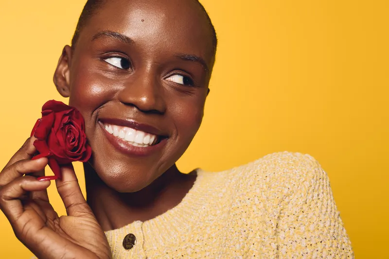 Happy African American woman with dark skin holding red rose near face smiles looking sideways. Yellow background and cozy sweater create warm and bright mood for beauty and joy concept.