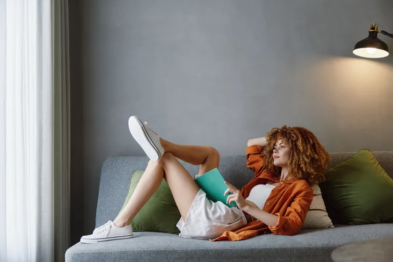 Красивая молодая женщина с кудрявыми волосами в оранжевой рубашке наслаждается досугом на диване с зеленой бутылкой воды