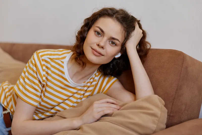 A relaxed young woman rests on a beige sofa at home, head tilted on a pillow with a gentle smile, expressing calm, comfort, and everyday self care in a cozy living space.