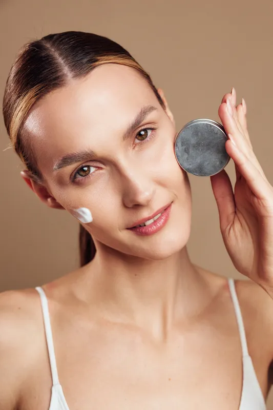 A young woman with clear skin applies cream to her face while holding a cosmetic jar. The close-up portrait shows skincare routine and natural beauty concept.