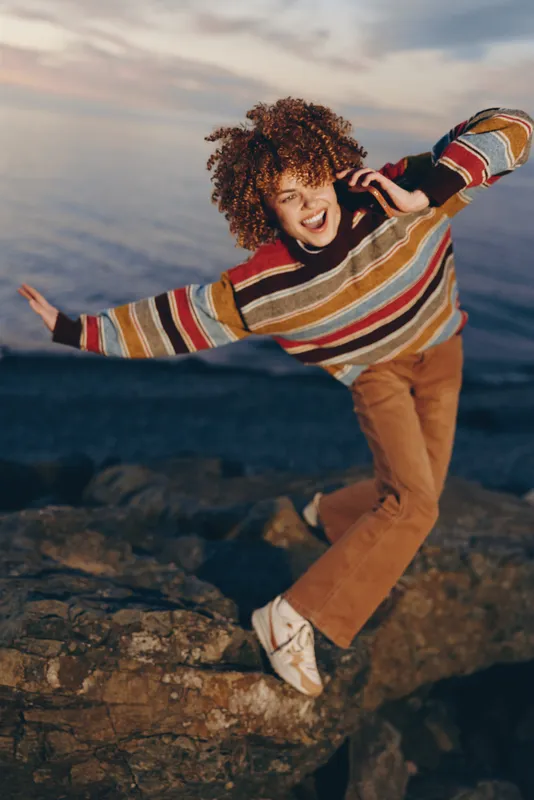 Joyful woman lifestyle on seaside rocks by the ocean, curly hair lifted in wind, striped sweater casual pants, stretching at sunset, outdoor portrait with waves behind, natural light