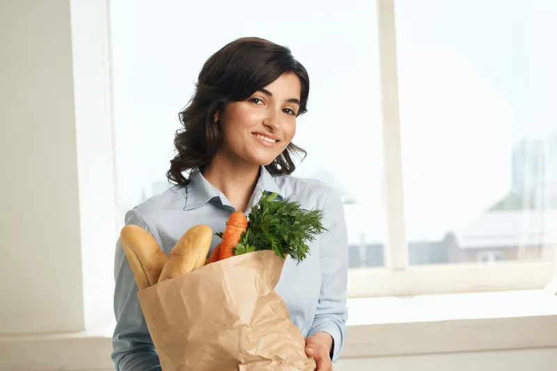 Весёлая брюнетка с пакетом продуктов, полным овощей, совершает покупки здоровой пищи
