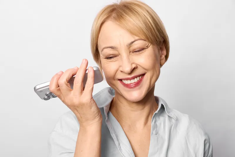 Woman smiling with smartphone making phone call, eyes closed, soft studio lighting on plain background. Communication technology supports wellness and everyday life.