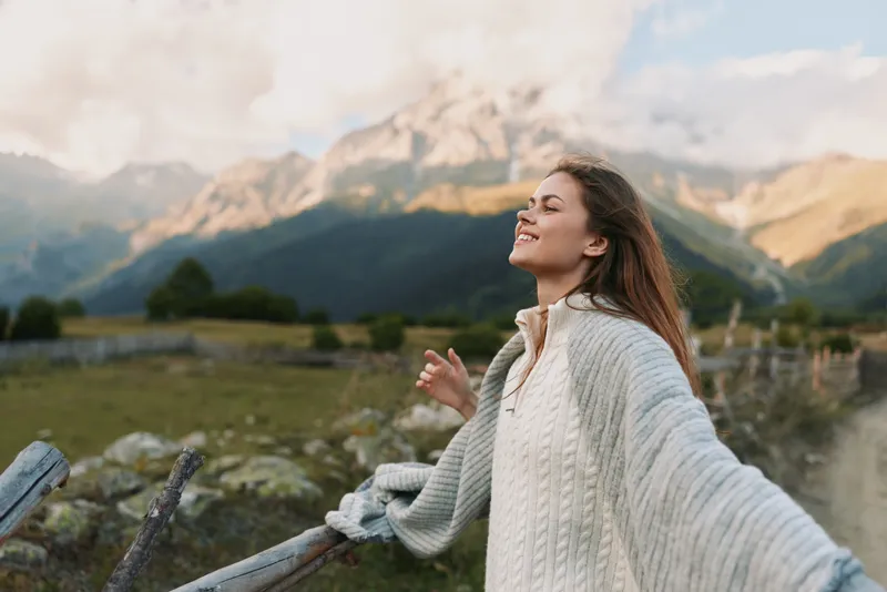 A woman stands outdoors on a sunny mountainside, arms outstretched, smiling, wearing a cozy sweater. Bright landscape highlights freedom, happiness, and a carefree lifestyle.