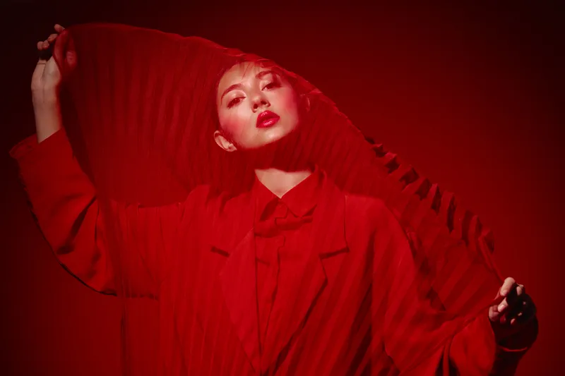 Fashion model in red coat posing with textured red fabric over face and intense makeup under red studio lighting. Concept of beauty creative fashion editorial.