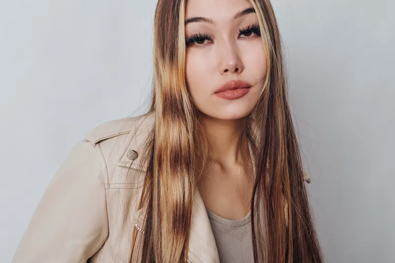 Young woman with long straight hair and natural makeup wearing beige jacket posing against plain background looking confident and stylish portrait.