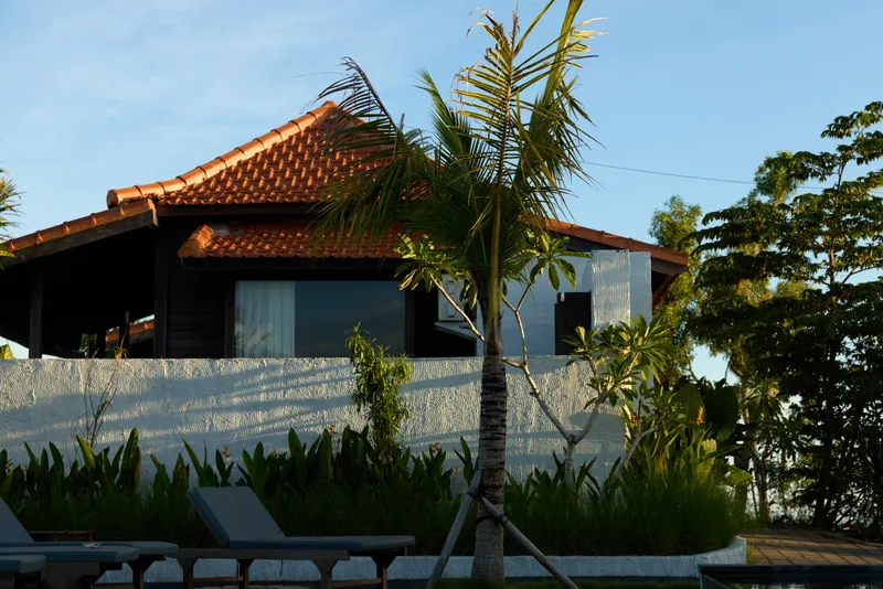 A tropical villa with a red roof, white wall, and palm trees invites outdoor relaxation. Sunlit deck chairs evoke a peaceful lifestyle getaway in a coastal setting.