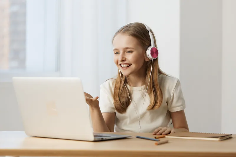 Young girl wearing headphones, smiling while engaged with her laptop at home, showcasing online learning in a bright and cheerful environment
