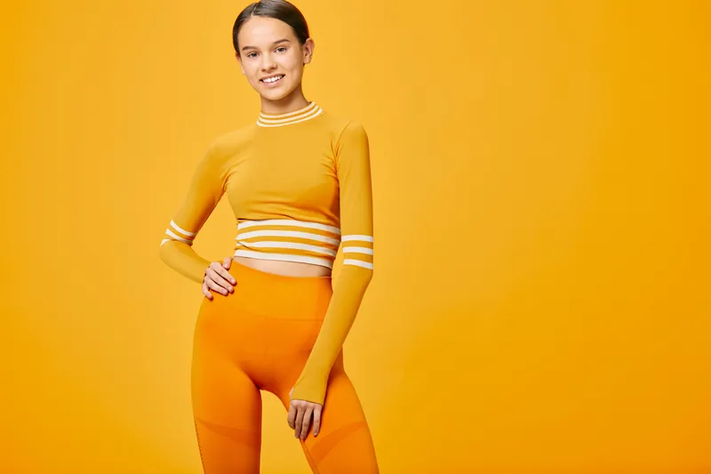 Fitness-minded woman poses in a vibrant orange athletic set against a bright yellow studio backdrop, showcasing strength, balance and confidence in a dynamic workout vibe.