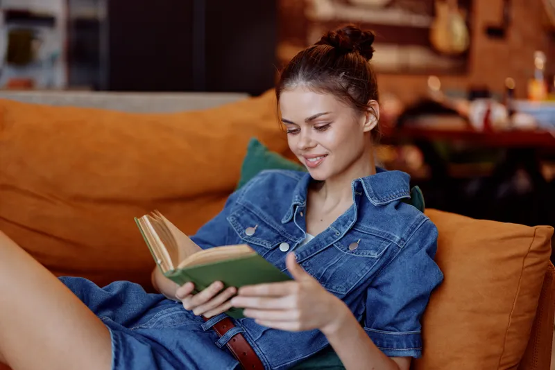 Женщина в джинсовой рубашке сидит на диване и читает книгу