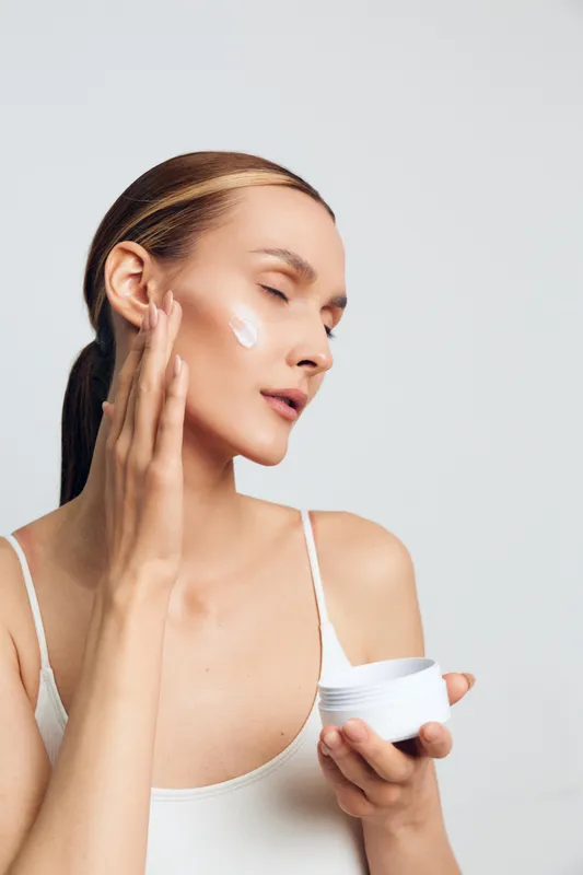 Young woman applying cream on her face with closed eyes skincare routine smooth skin natural beauty white background minimal makeup wellness