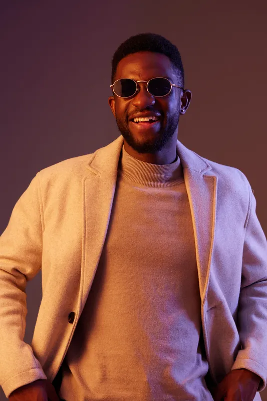 Stylish young man with sunglasses and a beige coat, smiling against a colorful background, representing confidence and modern fashion trends