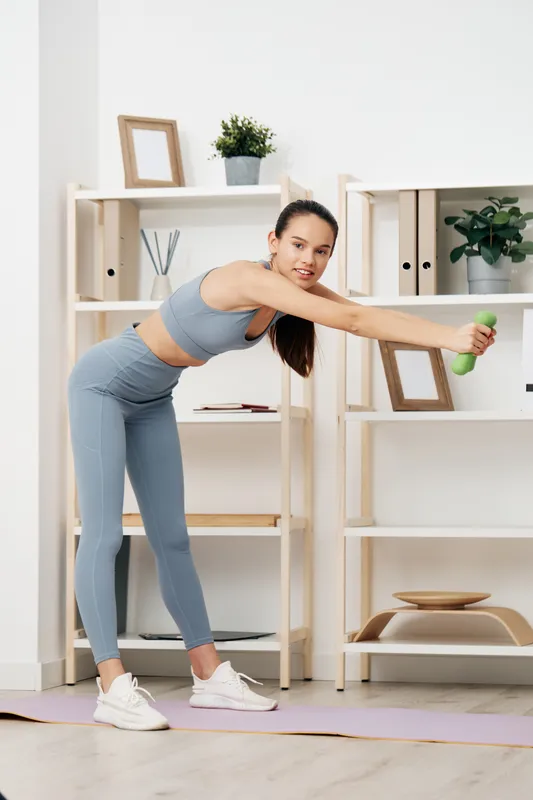 Woman fitness workout at home: young woman in matching blue activewear performs a side stretch holding a small green dumbbell on a yoga mat with wooden shelving and plants.