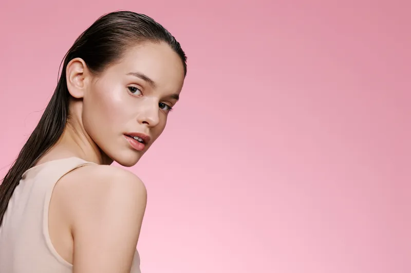 Young woman with wet hair looking back over her shoulder, soft pink background, nude dress, showcasing natural beauty and confidence, ideal for beauty and skincare concepts