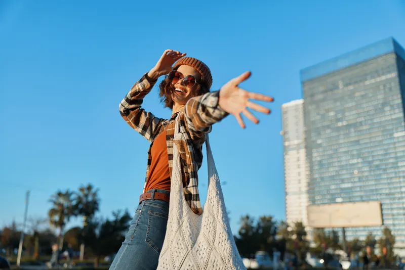 Woman smiling and reaching camera in urban park at golden hour, candid lifestyle portrait with authenticity and golden hour glow, mindful living vibe and emotional storytelling.