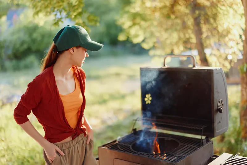 Young woman enjoying a sunny day grilling outdoors, wearing casual summer clothes and a cap, surrounded by greenery and warm sunlight