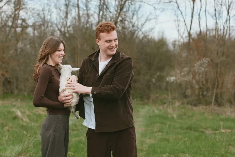 A lifestyle scene shows a couple outdoors with a goat kid, smiling and sharing warmth, affection, and joy in a natural rural setting, portraying bonding and animal companionship in daily life.