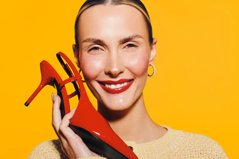 Smiling woman with red lipstick holds red high heel shoe near face on bright yellow background. Concept of fashion, style, beauty, and confident female model with vibrant colors.