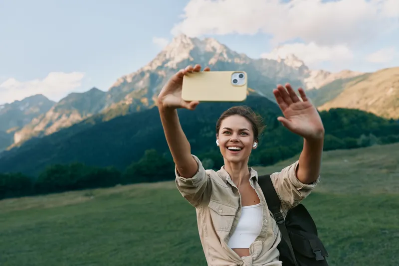Cheerful woman outdoors taking a selfie in a mountain meadow, bright smile, casual outfit, sunny day, sense of adventure, natural colors, scenic backdrop amid lush greens