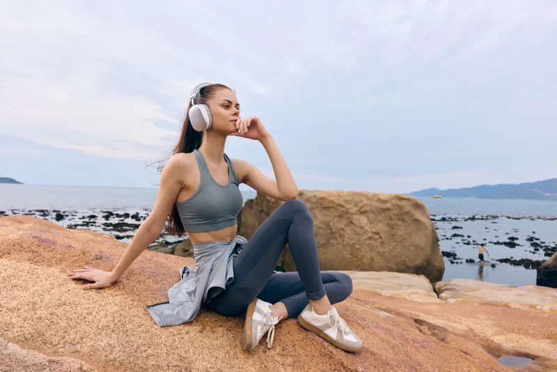 A fit young woman relaxing by the seaside, wearing sportswear and headphones, contemplating while enjoying the tranquil coastal landscape