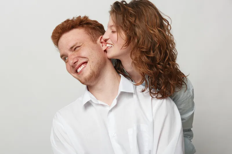 Happy young couple with curly hair and red hair share a joyful moment, woman kissing man on cheek. Studio portrait on white background, concept of love, affection, and relationship.