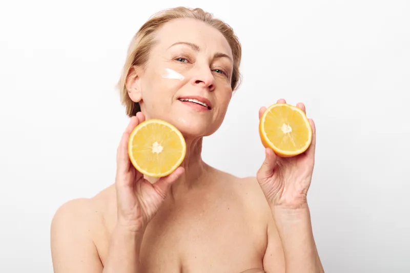Mature woman with short hair holds two halves of fresh orange close to face with cream on cheek. Concept of natural skincare, healthy lifestyle, and organic beauty treatment on white background.