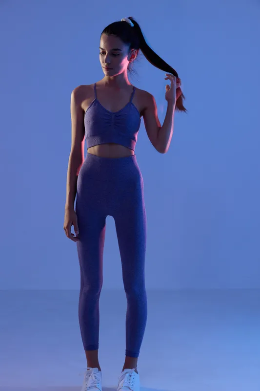 woman sport fitness dressed in purple activewear standing with a ponytail under blue studio lighting, posture showing calm focus and athletic form