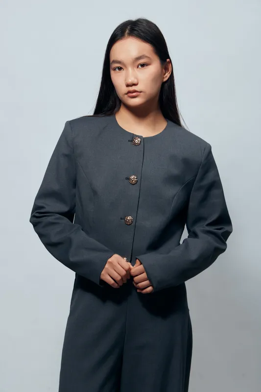 Young woman posing in dark gray tailored suit with buttoned jacket standing against plain light background. Fashion portrait with serious expression and long straight black hair in studio.