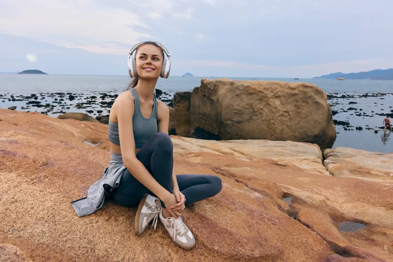 Young woman enjoying music with headphones, seated on rocky beach, calm expression, athletic attire, natural landscape, summer vibe, lifestyle concept