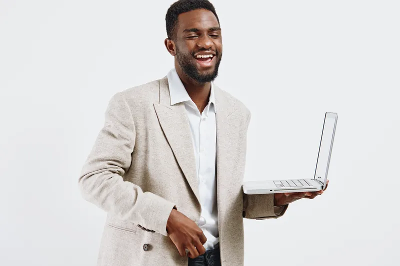 Smiling man wearing beige blazer and white shirt holding open laptop computer in hand, looking friendly, confident, and professional isolated on plain white background, studio portrait with modern bus