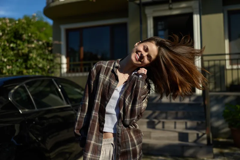 Smiling woman with flowing hair wearing a plaid shirt outdoors, enjoying sunlight. Casual lifestyle moment capturing happiness, confidence, and everyday style