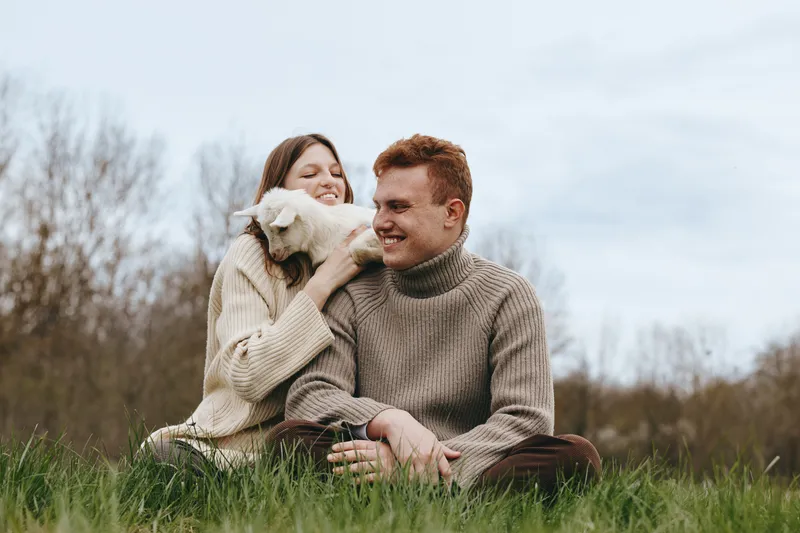 Lifestyle moment with a couple in cozy sweaters outdoors, sharing affection with a playful goat kid, sunlit grass and soft breeze, natural colors, peaceful countryside background