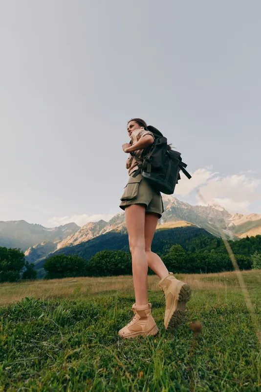 Cheerful woman hiking outdoors, backpack on, sunlit meadow with distant mountains, casual outfit, relaxed posture, natural colors, sense of adventure, fresh air, open landscape