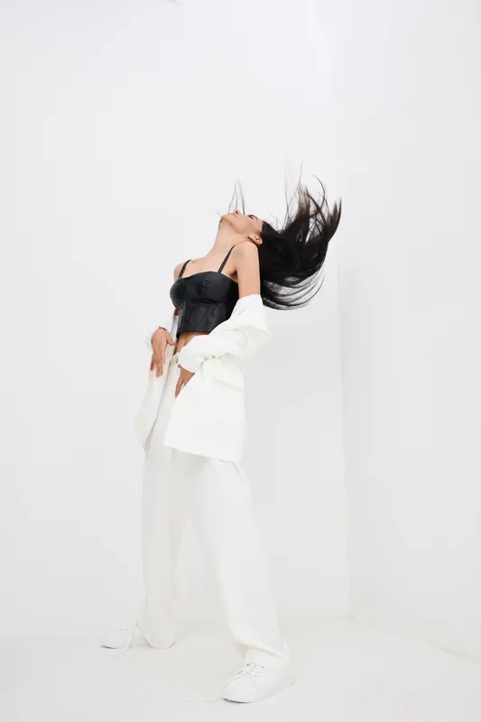 Woman with long dark hair in motion wearing white pants and jacket with black corset top, captured against a minimalistic white background in dynamic pose.