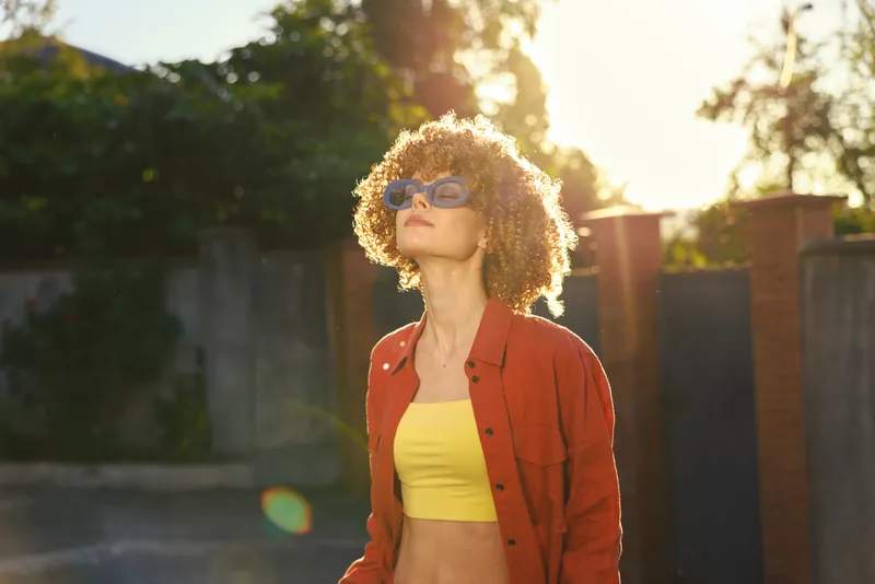 Smiling woman with curly hair wearing a yellow crop top and red jacket outdoors, backlit by sun. Casual fashion lifestyle shot with warm, relaxed mood and natural glow that conveys ease