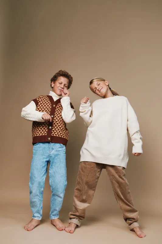 Two playful children pose in cozy autumn outfits, standing barefoot against a neutral backdrop, exuding cheerful energy, friendly charm, and a carefree, stylish mood.