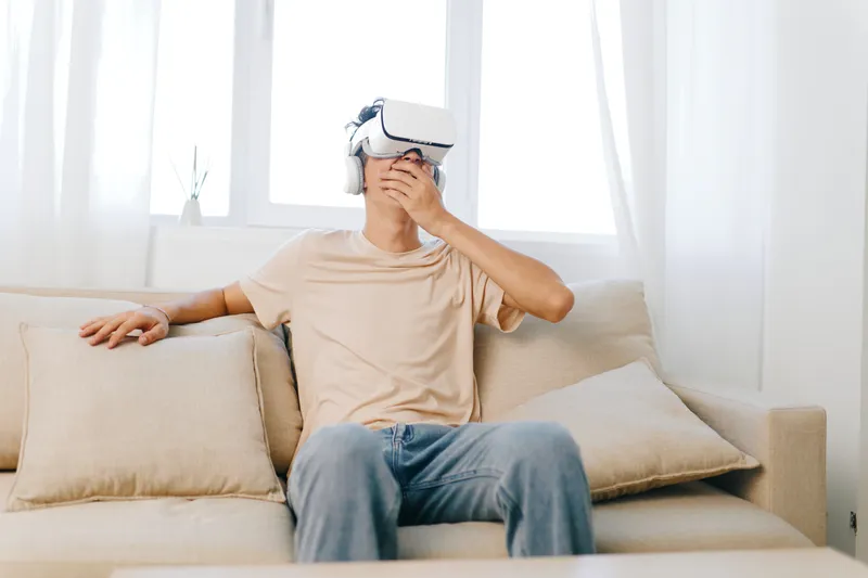 Молодой человек наслаждается опытом виртуальной реальности: он в шлеме VR сидит на мягком бежевом диване