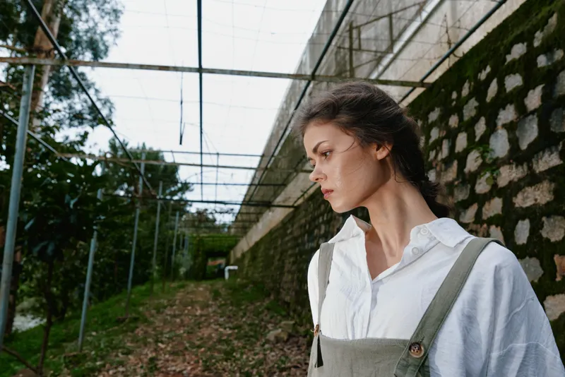 Young woman in overalls, showcasing a thoughtful expression, standing in an outdoor garden space with greenery and rustic walls, conveying a sense of serenity and nature