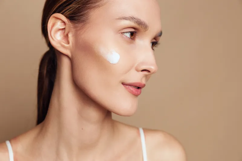 A young woman with clear skin applies cream on her cheek, looking to the side against a neutral background. Concept for skincare, beauty routine, and natural complexion.