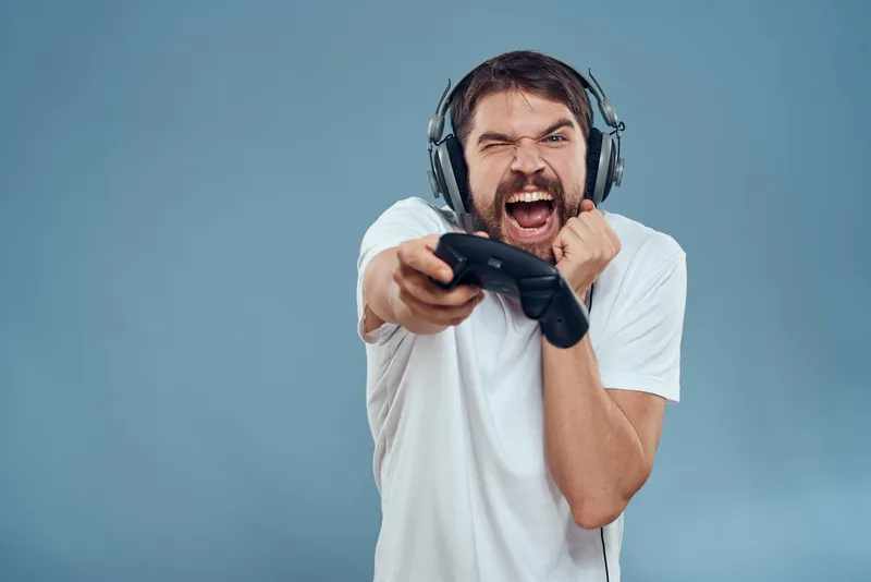 Мужчина в наушниках с контроллером в руках играет на игровой консоли, эмоции, образ жизни, синий фон, белая футболка