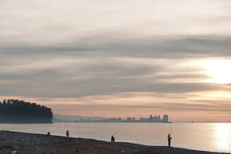 Beach sunset with silhouettes of fishermen along the shore, calm water and distant skyline, warm glow and golden light creating a tranquil coastal scene for travel and nature photography.
