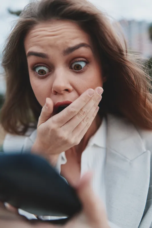 shocked woman with wide eyes covering mouth holding smartphone outdoors in business style clothes emotional financial crisis concept