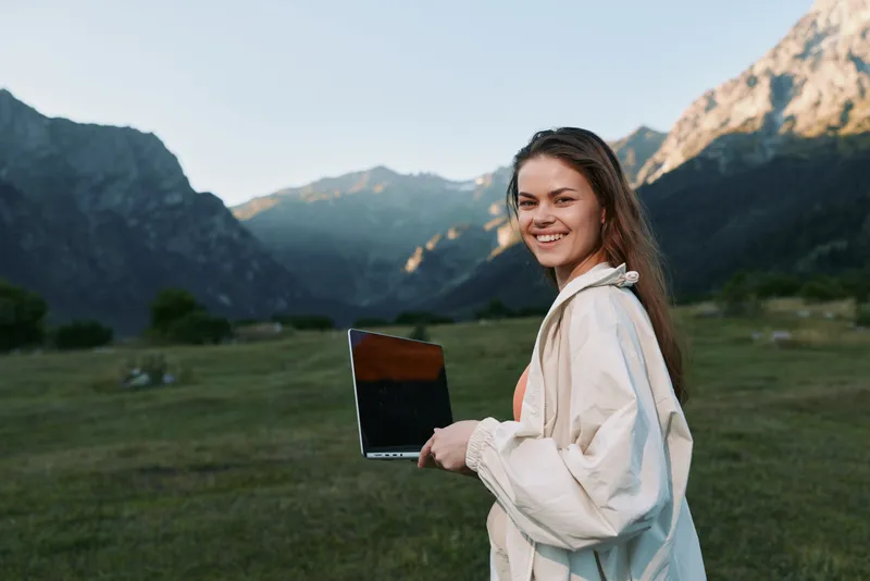 A cheerful professional woman stands outdoors in a mountain meadow, holding a laptop and smiling at the camera, projecting confidence, focus, and a positive modern work vibe.