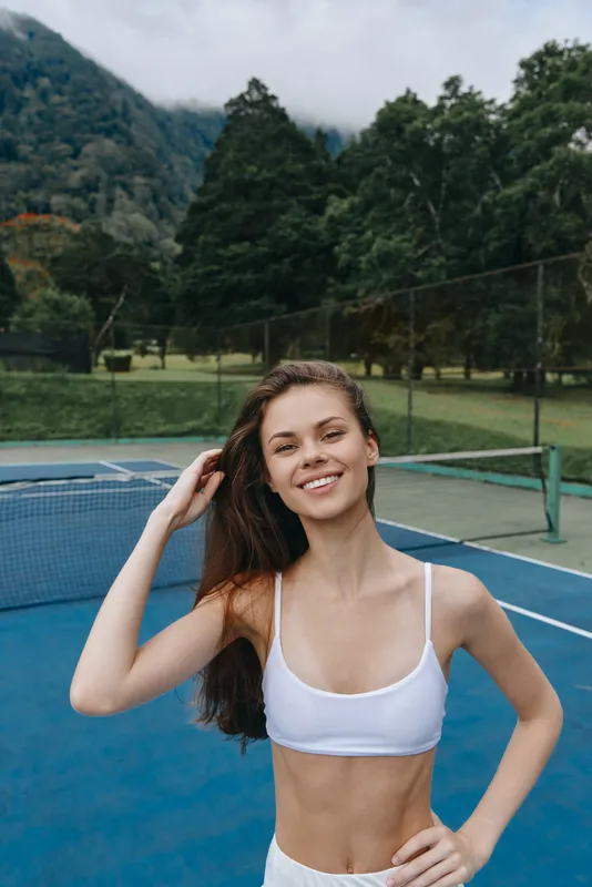 Athletic young woman smiling confidently on the tennis court, showcasing healthy lifestyle and fitness, dressed in sporty white clothing against a vibrant green backdrop