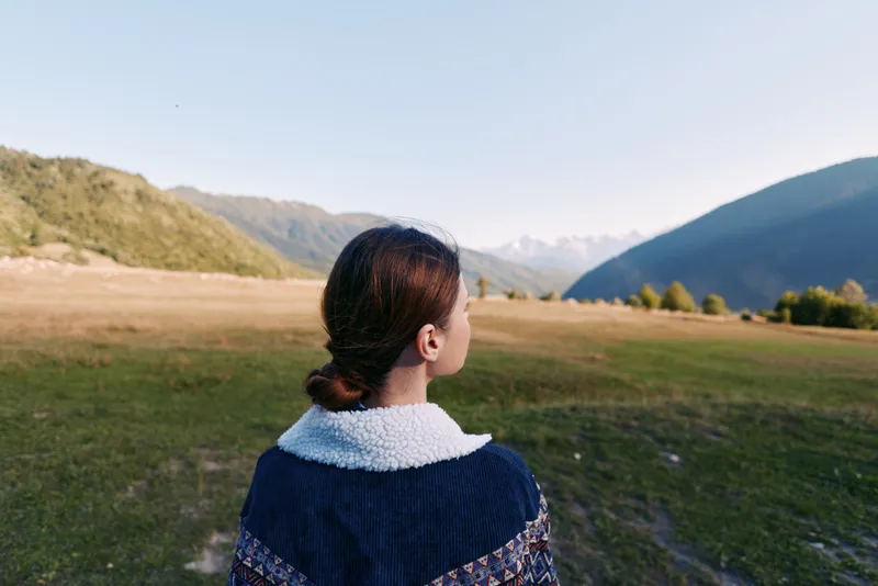 Woman back view looking at mountain valley and meadow landscape, nature travel scene with peaceful outdoors, horizon and distant peaks on a clear sky day