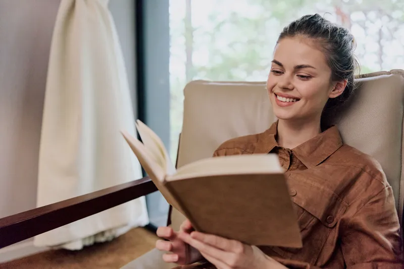 Улыбающаяся молодая женщина отдыхает, читая книгу в стильном интерьере, излучая комфорт и спокойствие